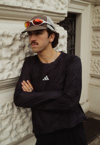 Mlad moški z brki, ki nosi kapo in rdeča odsevna sončna očala na kapi, stoji z prekriženimi rokami ob teksturirani kamniti steni.
