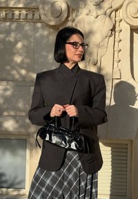 Femme aux cheveux noirs coupés en carré et portant des lunettes, vêtue d'un blazer sombre et d'une jupe à carreaux, tenant un sac à main en cuir noir devant un mur de pierre orné.
