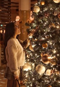 Árvore de Natal com agulhas verdes, adornada com ornamentos dourados, pretos e castanhos. Luzes brancas quentes realçam a atmosfera festiva.