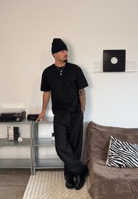 Svart oversized t-shirt och lösa svarta byxor med svarta stövlar. Mysig brun soffa med zebra-mönstrad kudde och växter i bakgrunden.