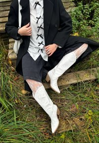 Vita knähöga cowboystövlar i texturerat skinn, med spetsiga tår och blockklack, kombinerade med en svart kappa och spetsstrumpbyxor över mörka shorts.