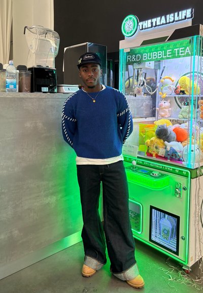 Suéter azul de punto con acentos blancos, gorra negra, camiseta blanca, jeans oscuros y zapatos beige, de pie cerca de una máquina de té de burbujas verde brillante.