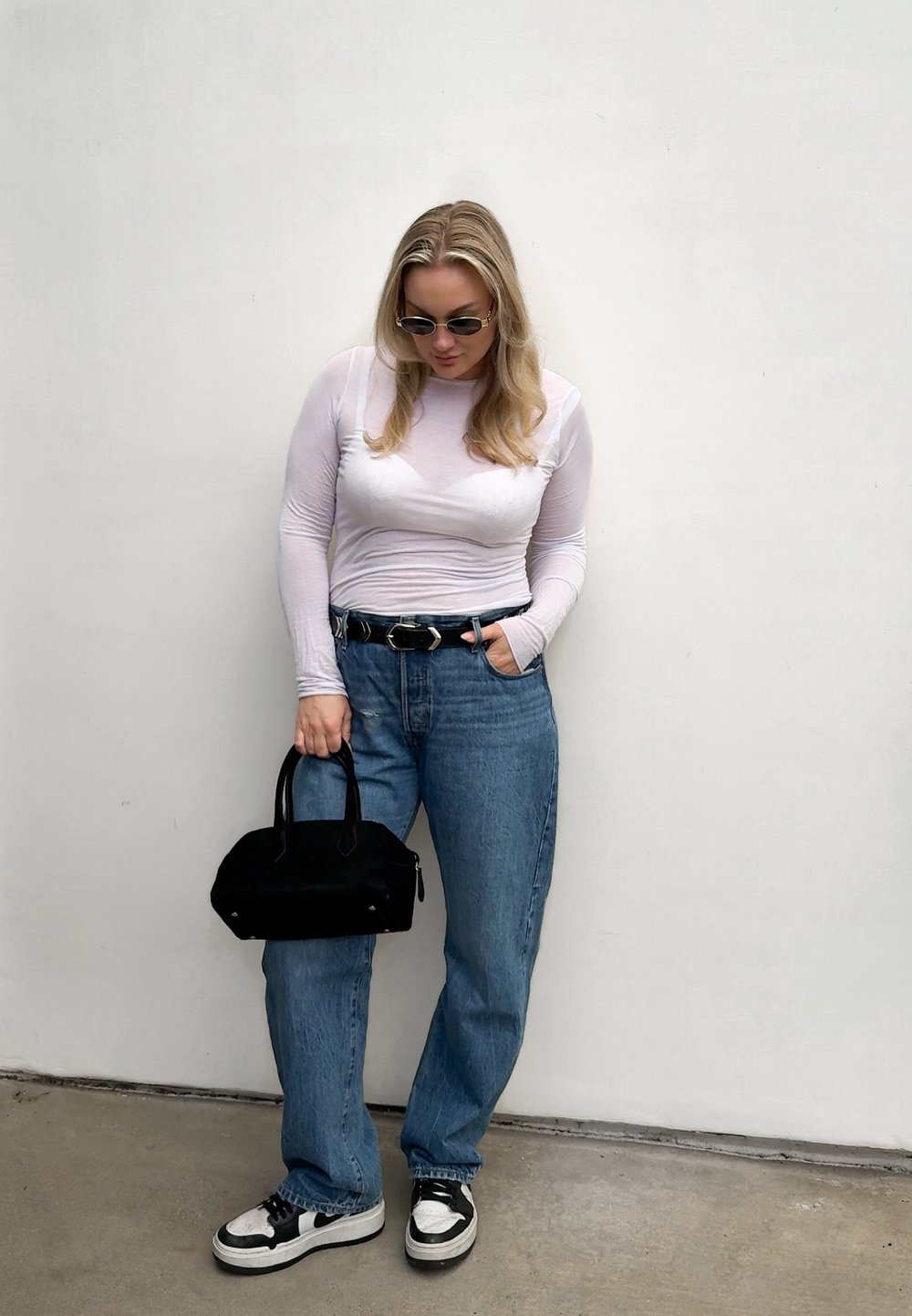 Femme portant un haut blanc à manches longues, un jean bleu, une ceinture noire et des baskets, tenant un sac à main noir, debout devant un mur blanc uni.