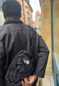 Svart North Face-ryggsäck med en böjd form, justerbart axelband, framfack och logotyp. Bärs över en svart jacka.