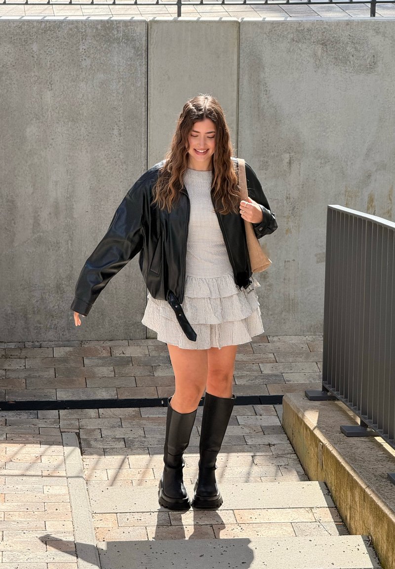 Νεαρή γυναίκα που φοράει φόρεμα με βολάν, μαύρο δερμάτινο μπουφάν και μπότες μέχρι το γόνατο, περπατά σε εξωτερικό χώρο σε πλακόστρωτη περιοχή δίπλα σε τσιμεντένιο τοίχο.