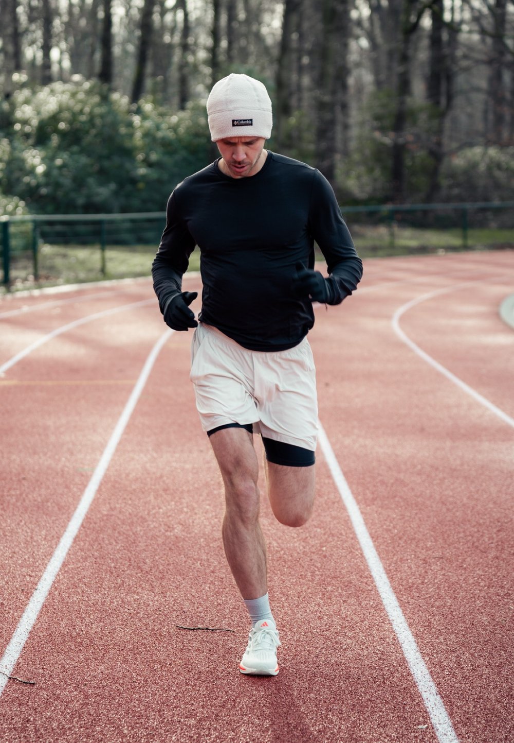 Urheilija juoksee punaisella radalla, päällään musta pitkähihainen paita, valkoiset shortsit ja valkoiset kengät oransseilla yksityiskohdilla. Pipossa ja hanskoissa.
