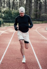 Urheilija juoksee punaisella radalla, päällään musta pitkähihainen paita, valkoiset shortsit ja valkoiset kengät oransseilla yksityiskohdilla. Pipossa ja hanskoissa.