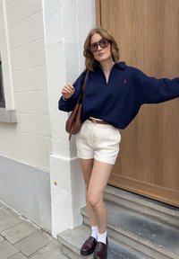 Marinskjorta med dragkedja, krämfärgade shorts, brunt bälte med guldfärgad hårdvara, mörka loafers och vita strumpor mot en trädd�örr och stentrappor.