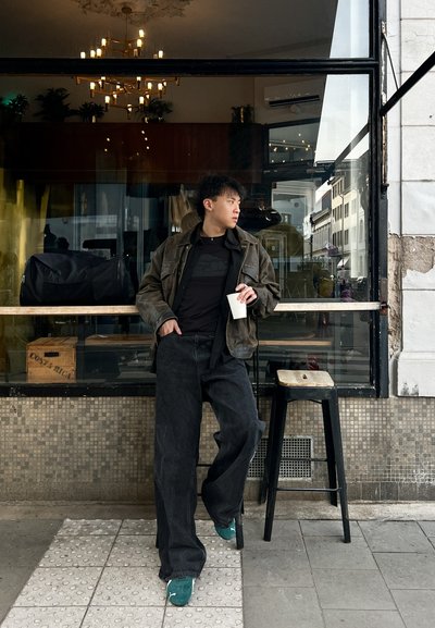 Chaqueta negra de gran tamaño y pantalones de mezclilla, combinados con zapatillas de deporte en color azul verdoso. De pie frente a un café, sosteniendo una taza blanca, junto a una bolsa negra.