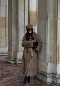 Manteau en fourrure marron avec un chapeau assorti, les deux présentant une texture douce. Des gants noirs et des bottes noires à talons hauts complètent la tenue devant des piliers en pierre.