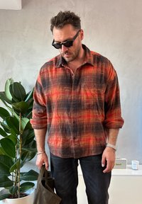 Muž s čiernymi slnečnými okuliarmi a červeno-čiernou károvanou košeľou, ktorý drží tmavú tašku, stojaci pri vysokom zelenom rastline v minimalistickej miestnosti.