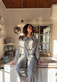 Set di pigiama a righe in azzurro chiaro e bianco, con colletto arricciato e dettagli a fiocco. Circondato da stoviglie e luce solare calda.