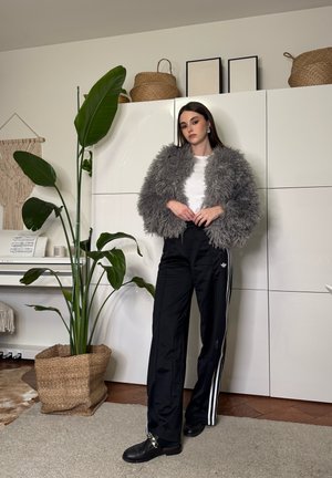 Mujer de pie en el interior, vestida con una chaqueta gris y peluda, camiseta blanca, pantalones deportivos negros de Adidas y zapatos negros, junto a una planta alta y verde en una cesta tejida.