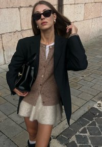 Sort blazer over en brun cardigan med knapper, cremet fuzzy nederdel og sort handbag. Runde guld øreringe og sorte solbriller fuldender looket.