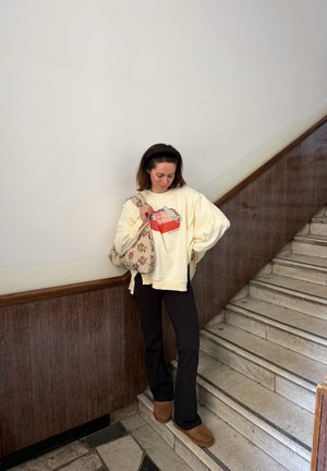 Mujer de pie en escaleras de mármol, vistiendo sudadera crema con diseño rojo, pantalones negros, zapatos marrones y llevando un bolso de hombro con estampado floral.