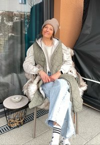 Καπιτονέ μακρύ γιλέκο σε ελαιόχρωμο πάνω από ένα ανοιχτό πουλόβερ, συνδυασμένο με ανοιχτό μπλε τζιν με φθορές. Τα αξεσουάρ περιλαμβάνουν ένα μπεζ σκουφάκι και λευκά sneakers.