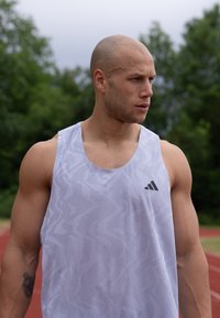 Hvit atletisk tanktopp med et mønstret tekstur, med en svart logo på brystet. Stoffet ser lett og pustende ut.