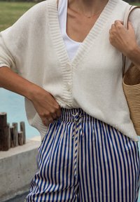 Cardigan in maglia crema con maniche corte e chiusura a bottoni, abbinato a pantaloni a vita alta a righe blu e beige e una borsa intrecciata.