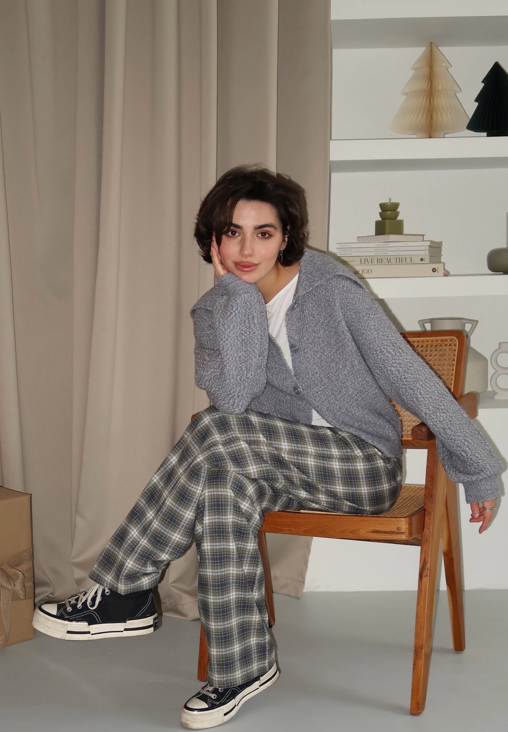 Cardigã cinza de malha, camiseta branca e calças largas em padrão xadrez nas tonalidades verde e azul. Ténis pretos com solas brancas. Cadeira de madeira, fundo neutro.