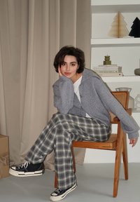 Cardigã cinza de malha, camiseta branca e calças largas em padrão xadrez nas tonalidades verde e azul. Ténis pretos com solas brancas. Cadeira de madeira, fundo neutro.