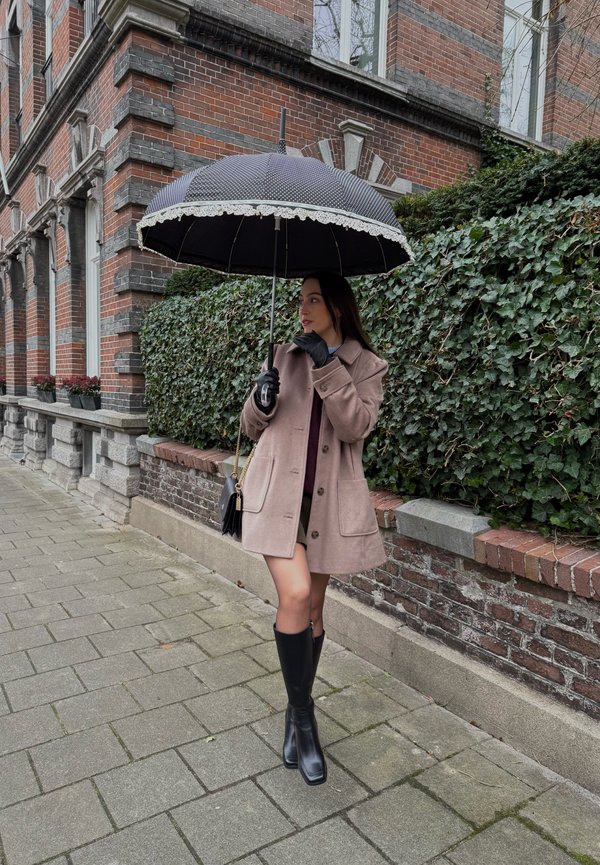 Vrouw in beige jas en zwarte laarzen die een zwarte paraplu met kanten rand vasthoudt, staand op het trottoir naast een bakstenen gebouw en een met klimop bedekte muur.