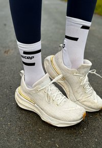 Bijele sportske tenisice s teksturiranom gornjom površinom, zlatnim detaljima na potplatu, uz bijele čarape s crnim grafičkim dizajnom. Noge u tamnoplavim tregersicama.