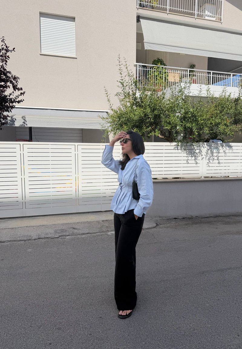 Femme portant des lunettes de soleil, un chemisier blanc et un pantalon noir, debout dans une rue à côté d'une clôture blanche et d'un bâtiment résidentiel avec verdure.