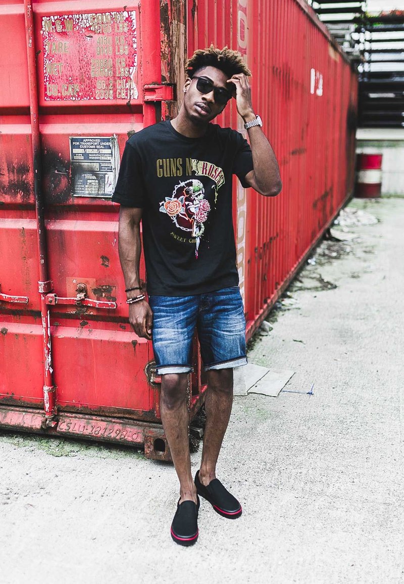 Black graphic t-shirt featuring 'GUNS N' ROSES' print; blue denim shorts; black slip-on shoes with red accents; standing next to a red shipping container.