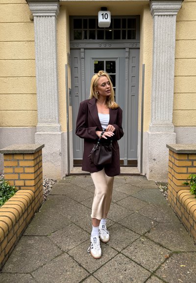 Blazer marrón oversized, top blanco y falda midi beige; sosteniendo un pequeño bolso negro. Combinado con zapatillas blancas y calcetines blancos. Fondo de pared de piedra.