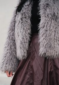 Veste grise en fausse fourrure à la texture moelleuse, portée sur un haut noir côtelé et une jupe bordeaux brillante, mettant en valeur la variété des matériaux et des couleurs.