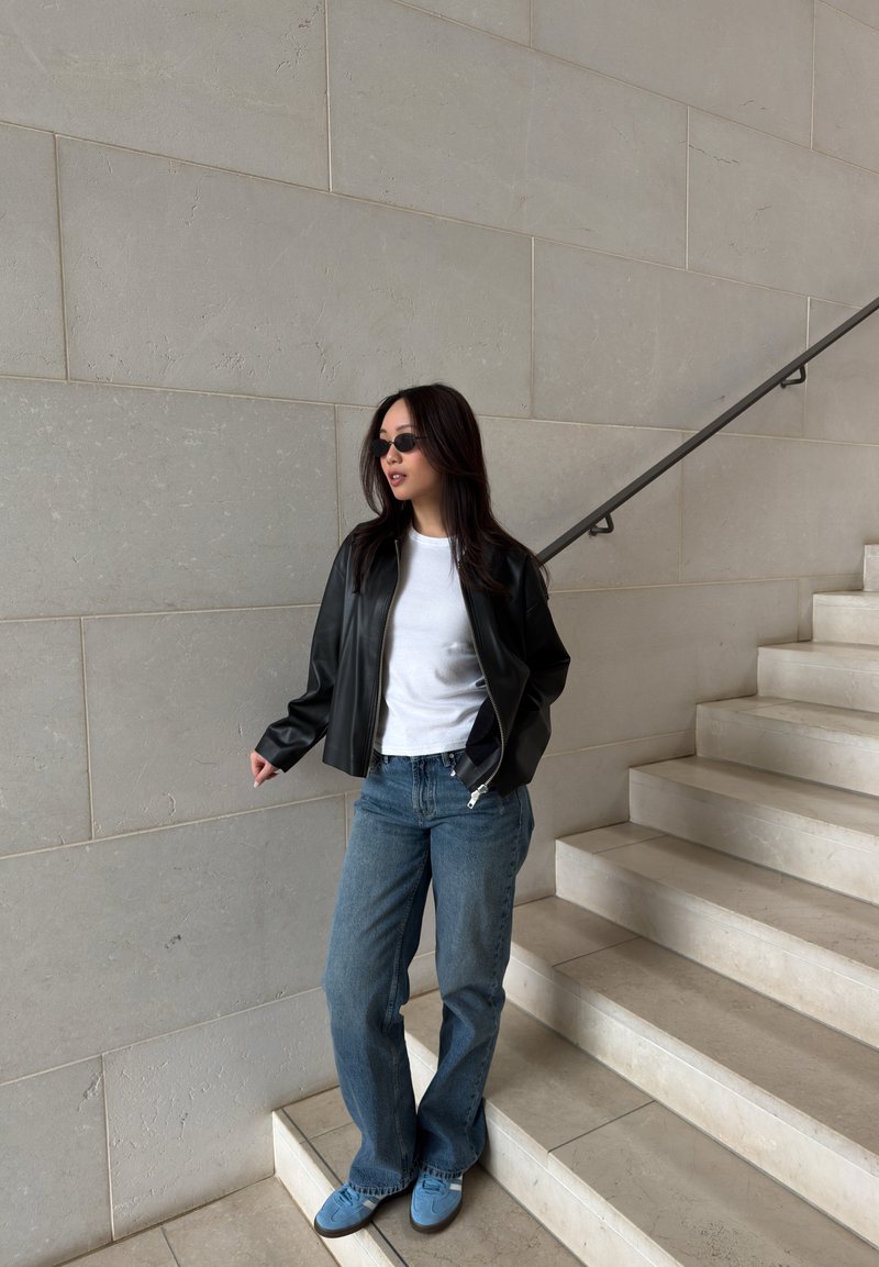Jeune femme portant des lunettes de soleil, une veste en cuir noire, une chemise blanche, un jean bleu et des baskets, se tient près d'un escalier et d'un mur en pierre beige.