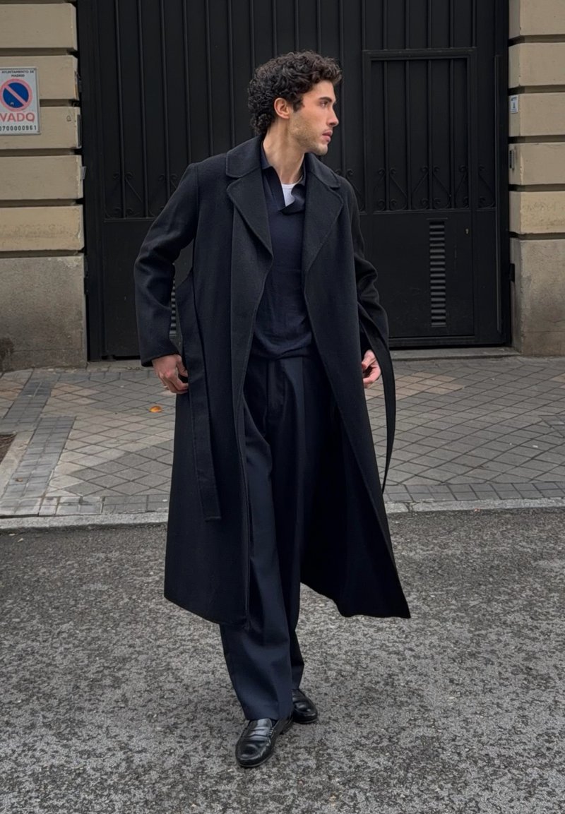 Homme aux cheveux bouclés portant un long manteau noir, un pantalon foncé et des chaussures noires, marchant sur un trottoir carrelé avec une grande porte noire derrière lui.