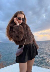 Femme portant des lunettes de soleil foncées, une veste en fourrure marron et une jupe noire, debout près de la mer au coucher du soleil sous un ciel nuageux.