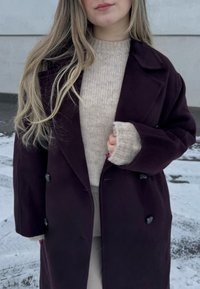 Άτομο που φοράει σκούρο μοβ παλτό πάνω από μια μπεζ πλεκτή μπλούζα, στέκεται έξω σε χιονισμένη επιφάνεια, κρατώντας το παλτό κλειστό με το ένα χέρι.