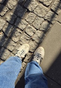 Lys grå sneakers med teksturert semsket skinn og mesh-overdel, med svart striper, sammen med lyseblå denimjeans på brosteinsbelagte fortau.