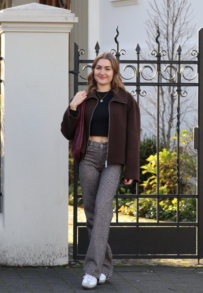 Chaqueta marrón de cremallera sobre un top corto negro, combinada con pantalones acampanados grises estampados y zapatillas blancas. Los accesorios incluyen un bolso de rafia.