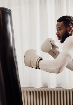 Hombre con camisa blanca de manga larga y guantes de boxeo golpea un saco de boxeo negro en una habitación con cortinas blancas.