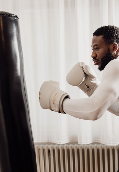 Hombre con camisa blanca de manga larga y guantes de boxeo golpea un saco de boxeo negro en una habitación con cortinas blancas.