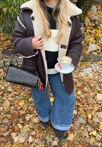 Brun frakke foret med fåreskind, sort-hvid stribet striktrøje, vidde, lige denimjeans, brune støvler, holder en kop kaffe med en lyserød makron.