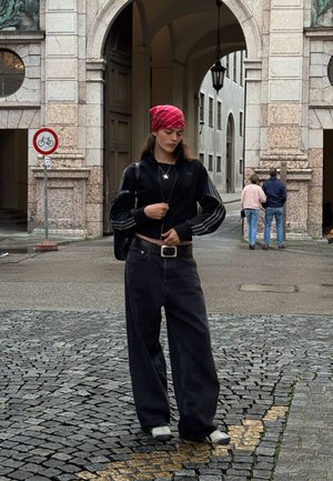 Juodas trumpas švarkas su trimis juostelėmis, laisvi juodi džinsai, raudonas languotas bandanas, sidabrinė žvaigždės pakabukas ir juodi sportbačiai ant akmenimis grįsto grindinio.