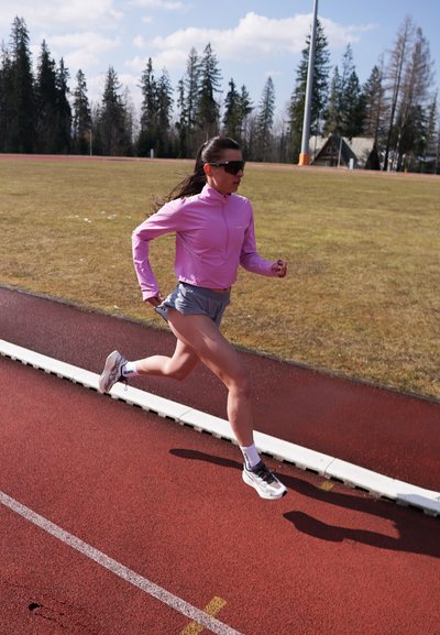 Mujer con chaqueta rosa, pantalones cortos grises y gafas de sol corriendo en una pista roja al aire libre con campo de césped y árboles al fondo.