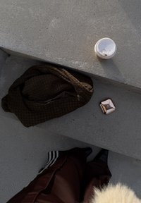 Borsa intrecciata marrone su cemento grigio con un piccolo portachiavi metallico e una tazza di caffè con coperchio nelle vicinanze. Pantaloni scuri con strisce bianche sul fondo.