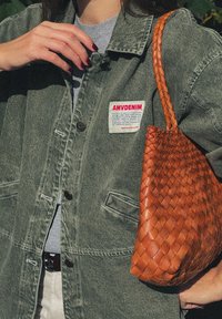 Giacca di jeans verde con chiusura a bottoni e toppa. Borsa in pelle intrecciata marrone con superficie testurizzata. T-shirt grigia sotto.