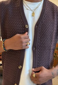 Person som bærer en brun teksturert cardigan over en hvit skjorte, gullkjeder, ringer, et blått og gullfarget perlekjede, og viser en tatovert underarm.