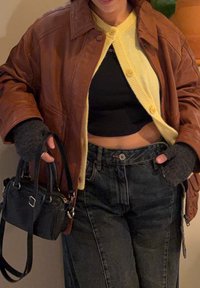 Personne portant une veste en cuir marron, un cardigan jaune, un top noir court et un jean foncé, tenant un sac à main noir avec des gants sans doigts.