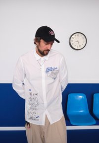 White button-up shirt featuring blue graphic designs and text, matched with beige trousers. A black cap with a pink logo is visible; blue chairs are in the background.