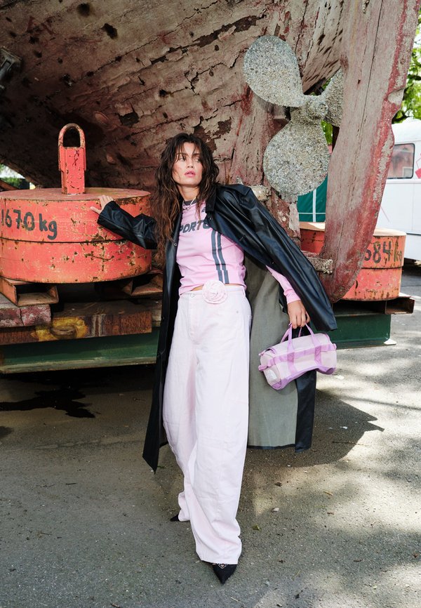 Vrouw in roze top, witte wijde broek en zwarte jas houdt een roze tas vast en leunt tegen grote roestige machine met metalen propeller buiten.