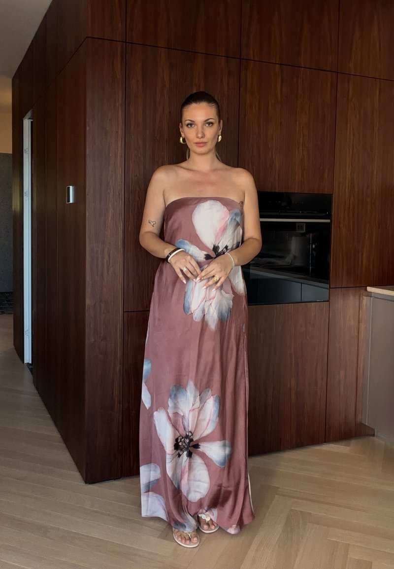 Femme debout à l'intérieur portant une robe mauve bustier avec de grands motifs floraux blancs et bleus, des boucles d'oreilles créoles dorées et des sandales.