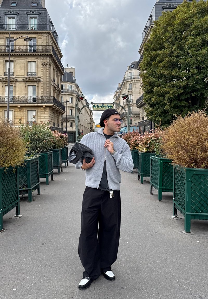 Pull gris zippé sur une chemise noire, pantalon noir large et chaussures noires avec des accents blancs. Entouré de verdure et d'architecture urbaine.