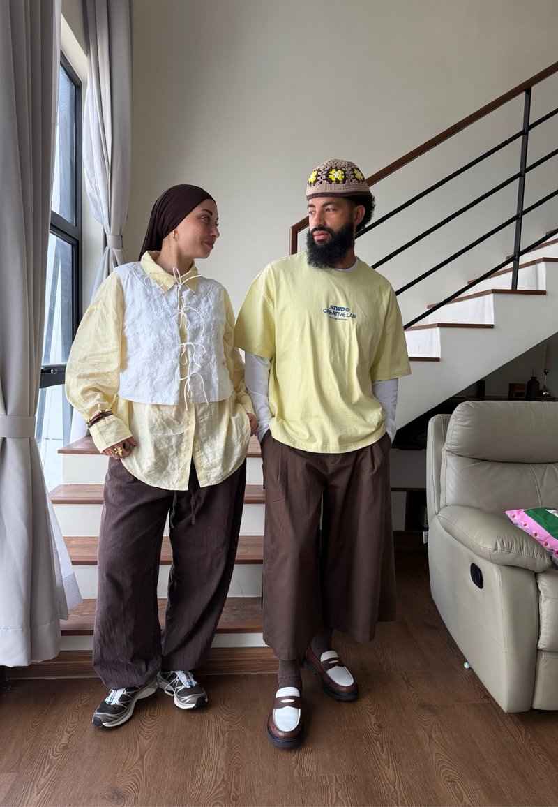 Deux personnes debout à l'intérieur près d'un escalier, portant des pantalons marron larges et des hauts jaune clair, se regardant dans un cadre décontracté.